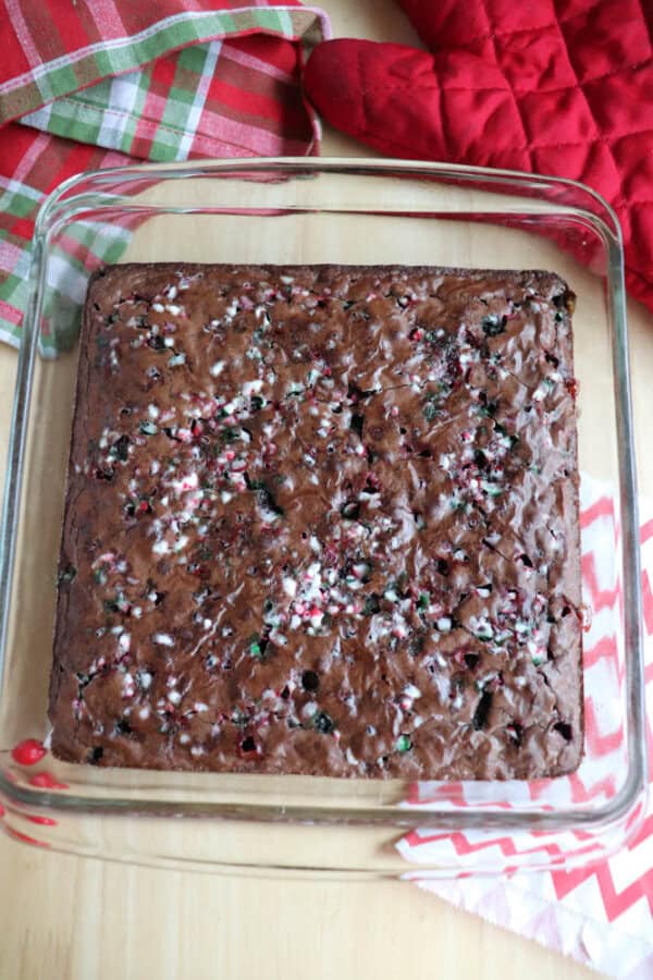 A square pan of freshly baked, festive brownies topped with crushed peppermint candies. The background includes a red oven mitt and colorful plaid and chevron-patterned cloths placed on a wooden table.
