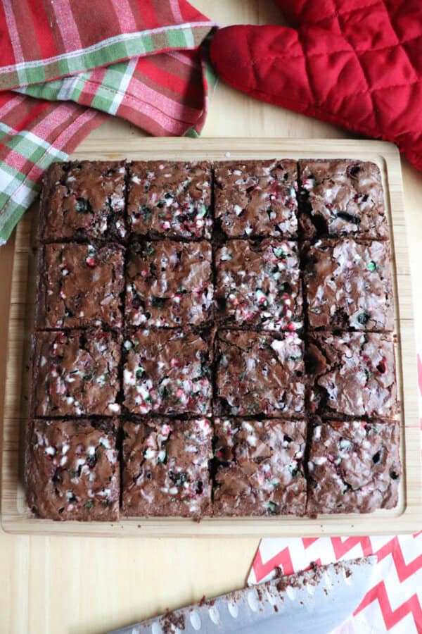 A freshly baked batch of brownies cut into squares on a wooden board. The brownies are topped with bits of crushed candy canes. Red and green plaid cloth, a red oven mitt, and a knife with chocolate crumbs are nearby.
