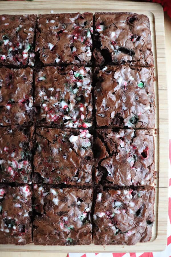 A close-up of a freshly baked batch of brownies, cut into 16 squares on a wooden board. The brownies are topped with crushed red and white peppermint candy pieces, adding a festive touch. The edges are slightly crisp, and the centers appear fudgy.