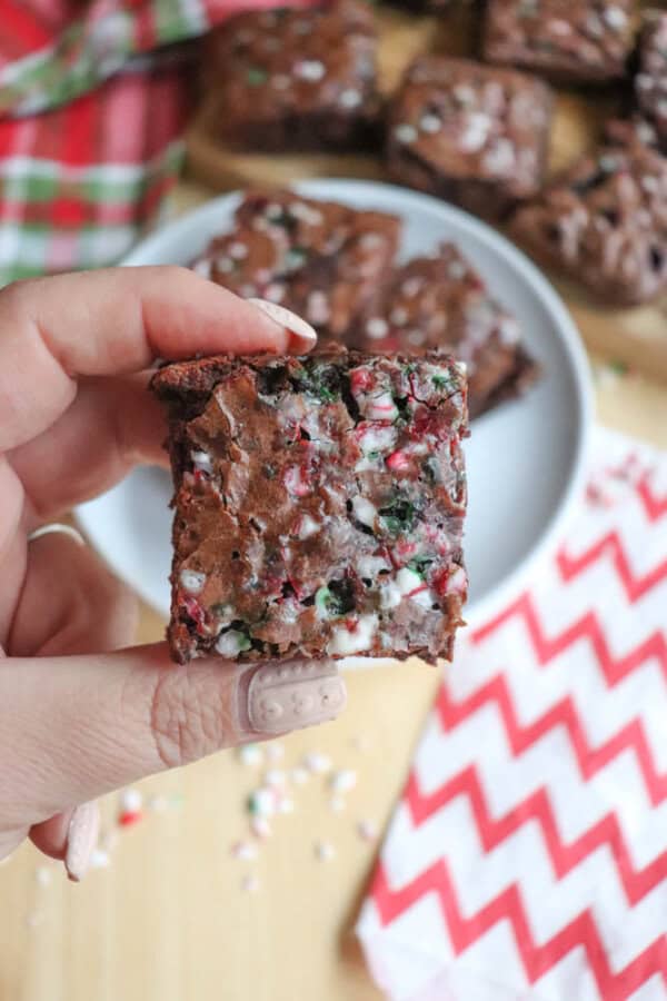 A hand holding a festive brownie with colorful sprinkles inside. The brownie has a crackled top and is held over a wooden surface with more brownies on a plate in the background, alongside a red and white chevron napkin.