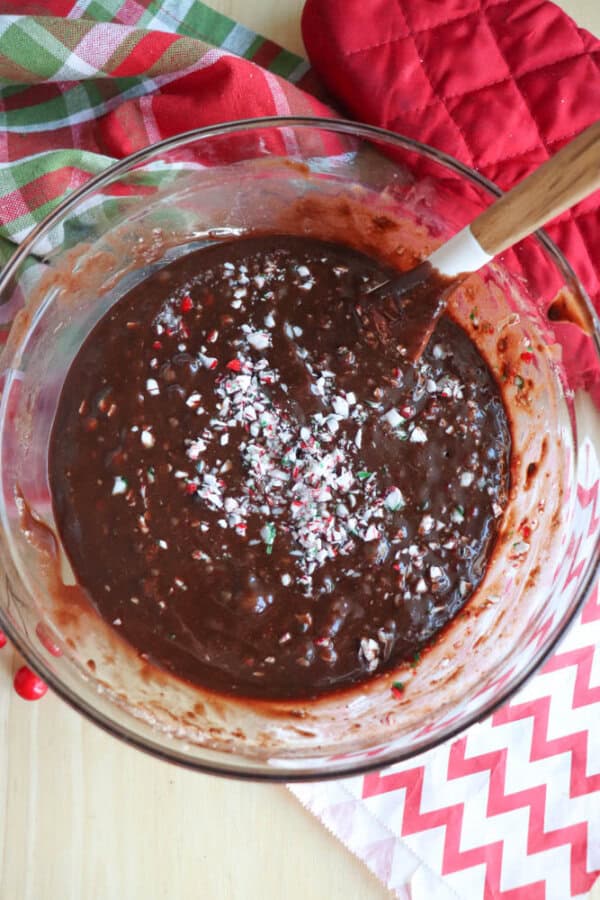 A glass bowl filled with rich chocolate batter topped with crushed peppermint pieces. A wooden spoon rests inside the bowl. Red oven mitts and a festive plaid cloth are nearby on a light-colored surface.