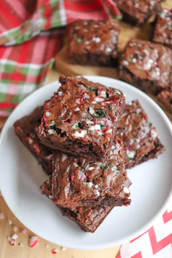 A plate of chocolate brownies topped with crushed candy canes sits on a wooden table. In the background, more brownies and a red and green plaid cloth are visible. The brownies have a festive, holiday appearance.