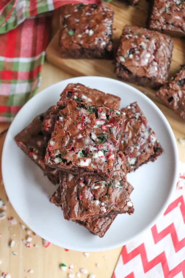 A plate with brownies topped with crushed peppermint candy. The brownies are stacked and placed on a festive red and green plaid cloth. Nearby, more brownies sit on a wooden board. Scattered candy pieces and a red zigzag-patterned napkin are visible.
