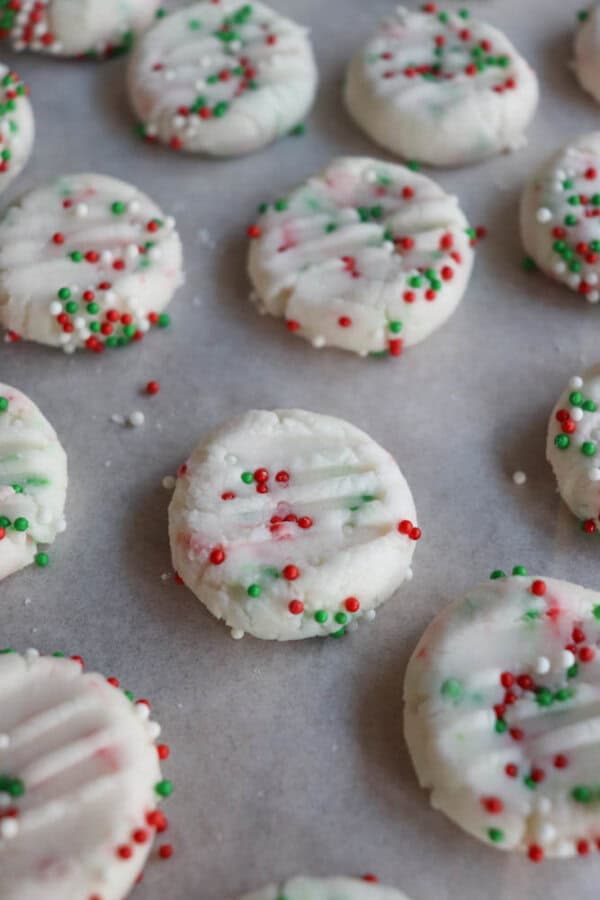 Round cookies with white frosting and red and green sprinkles are arranged on parchment paper. The sprinkles are evenly distributed across the cookiesโ surfaces.