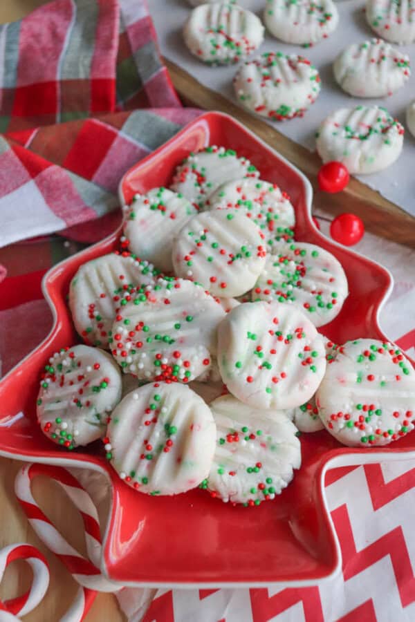 A red, tree-shaped plate filled with white cookies decorated with red and green sprinkles. A plaid cloth and more cookies are in the background, along with two red candy balls and a candy cane.