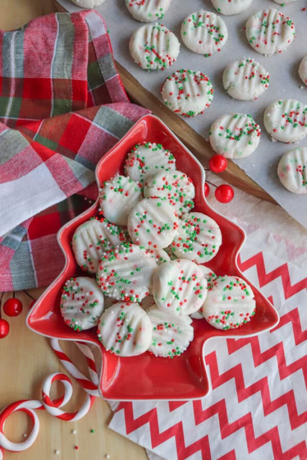 A Christmas-themed display with white chocolate-covered cookies topped with red and green sprinkles. The cookies are in a red, tree-shaped dish, and more are on a tray. A red and green plaid cloth and zigzag pattern napkin add festive accents.