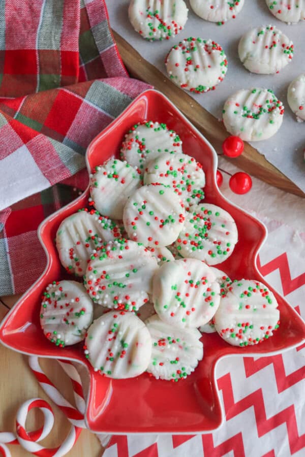 A tree-shaped red platter holds white chocolate-covered cookies decorated with red, green, and white sprinkles. Surrounding elements include a plaid cloth, more cookies on a tray, red candies, and candy canes on a chevron-patterned surface.