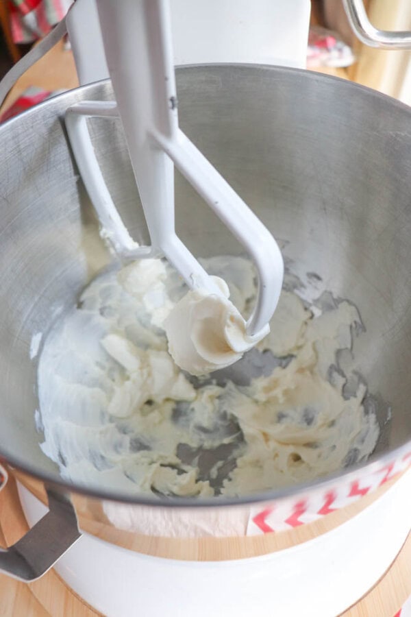 A mixer with a paddle attachment is partially filled with smooth, creamy white frosting in a metal mixing bowl. The mixer is on a wooden surface.