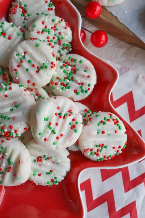 A festive plate of white cookies adorned with red, green, and white sprinkles. The cookies are arranged on a red tray shaped like a Christmas star. A zigzag-patterned tablecloth enhances the holiday theme.