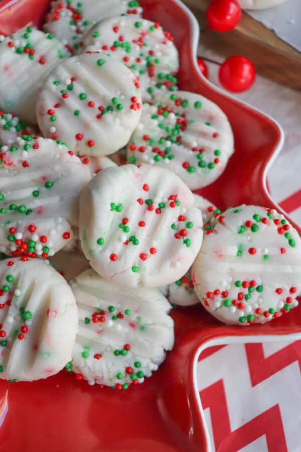 A festive plate of white cookies topped with red, green, and white sprinkles, arranged on a red star-shaped dish. The cookies have fork marks on top, enhancing their decorative appearance.