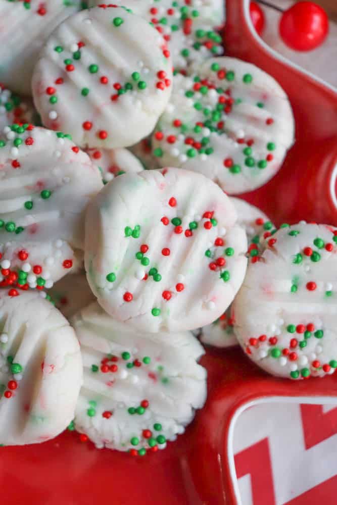 A stack of festive holiday cookies with white frosting, decorated with red, green, and white sprinkles, arranged on a red plate with a chevron pattern.