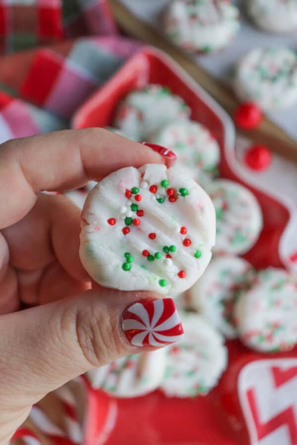 A hand with festive red and white nail art holds a white peppermint cookie decorated with red and green sprinkles. More cookies are visible in the blurred background, with a red and white striped cloth nearby.