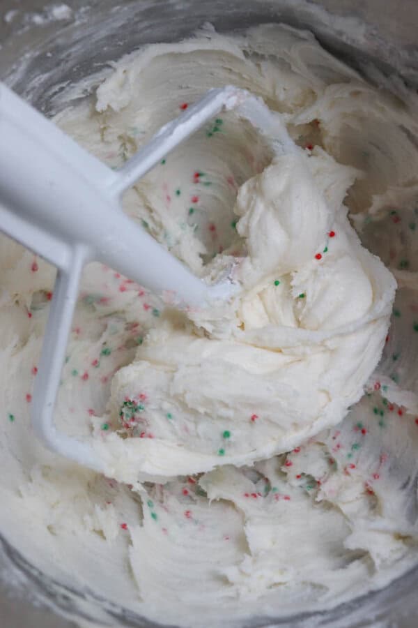Close-up of creamy white cake batter in a mixing bowl, speckled with red and green sprinkles, being mixed by a white paddle attachment.