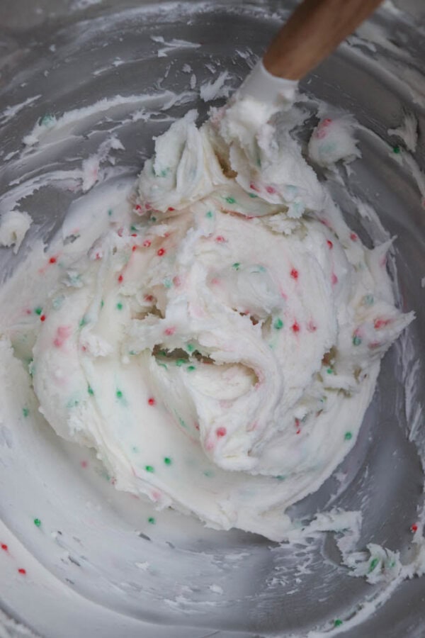 A bowl filled with white frosting mixed with small red and green sprinkles, being stirred with a spatula.