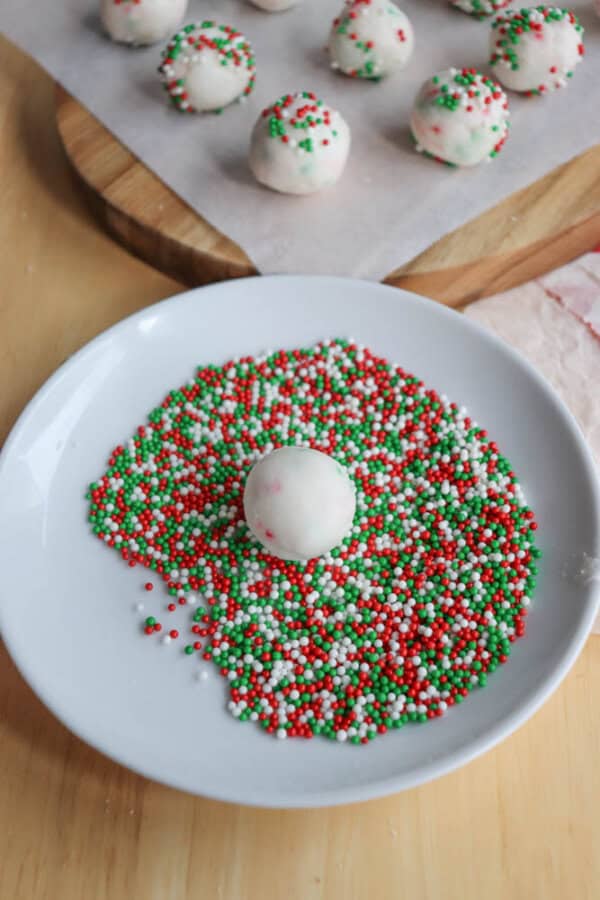 A white plate with red, green, and white sprinkles. A white truffle ball is in the center of the sprinkles. More truffle balls are in the background on parchment paper atop a wooden surface.