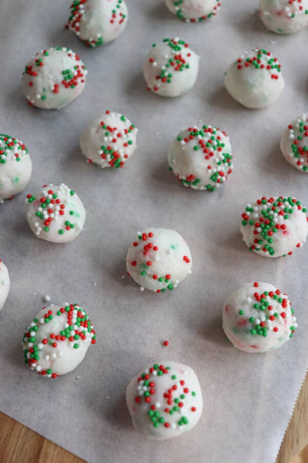 White-coated treats arranged on parchment paper, each topped with red, green, and white sprinkles, resembling festive holiday cake balls or cookies.