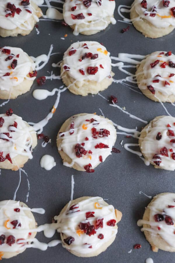 A tray of round cookies drizzled with white icing, topped with pieces of dried cranberries and orange zest. The cookies are evenly spaced on a dark baking sheet.