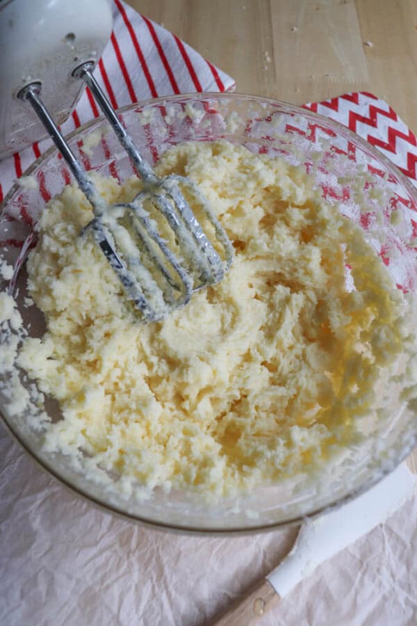 A glass bowl filled with mashed potatoes and a mixer with beaters resting in it. A red and white striped towel is underneath, and a white spatula is nearby on a wooden surface.