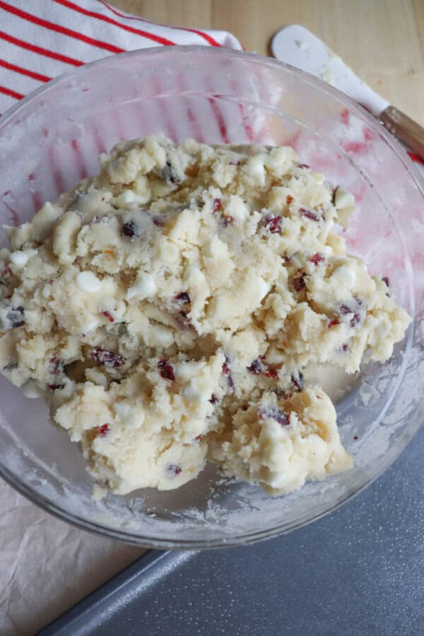 A glass bowl filled with cookie dough featuring white chocolate chips and dried cranberries. The bowl rests on a countertop next to a red striped kitchen towel and a spatula.