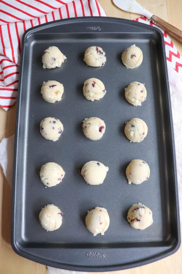 A baking sheet with evenly spaced scoops of cookie dough, ready to be baked. A red and white striped cloth is partially visible in the corner.