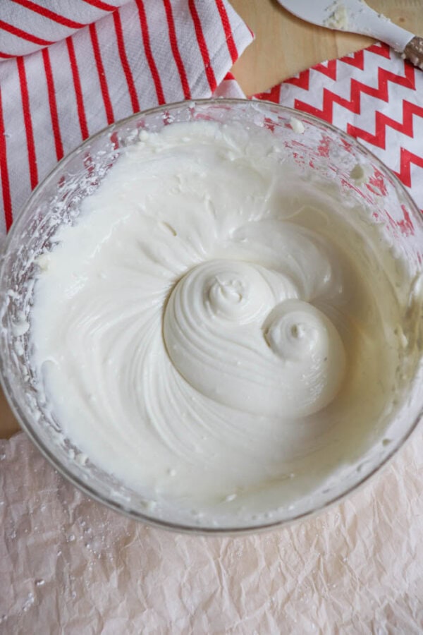 A glass bowl filled with smooth, creamy frosting sits on a table. The frosting has been whipped to a fluffy texture, and swirl patterns are visible on the surface. A red and white striped cloth is partially visible in the background.