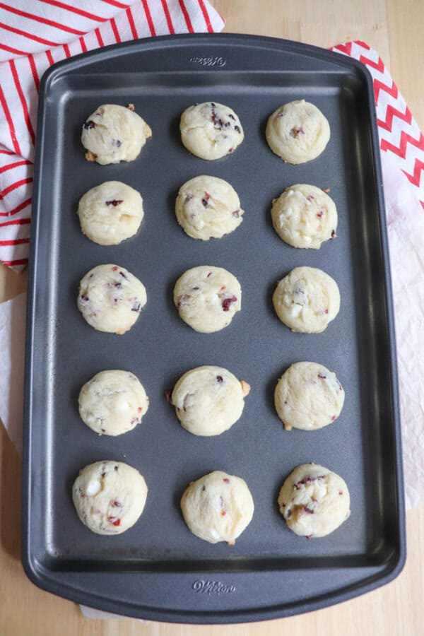 A baking tray holds sixteen freshly baked cookies with bits of dried fruit. The tray is on a wooden surface with a red and white striped cloth partially visible in the background.