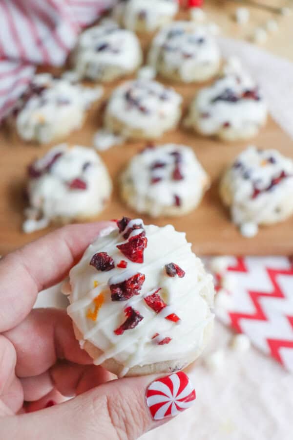 A hand holding a cookie topped with white icing and dried cranberries. More cookies are on a wooden board in the background. The person has a festive red and white peppermint design on their nail.