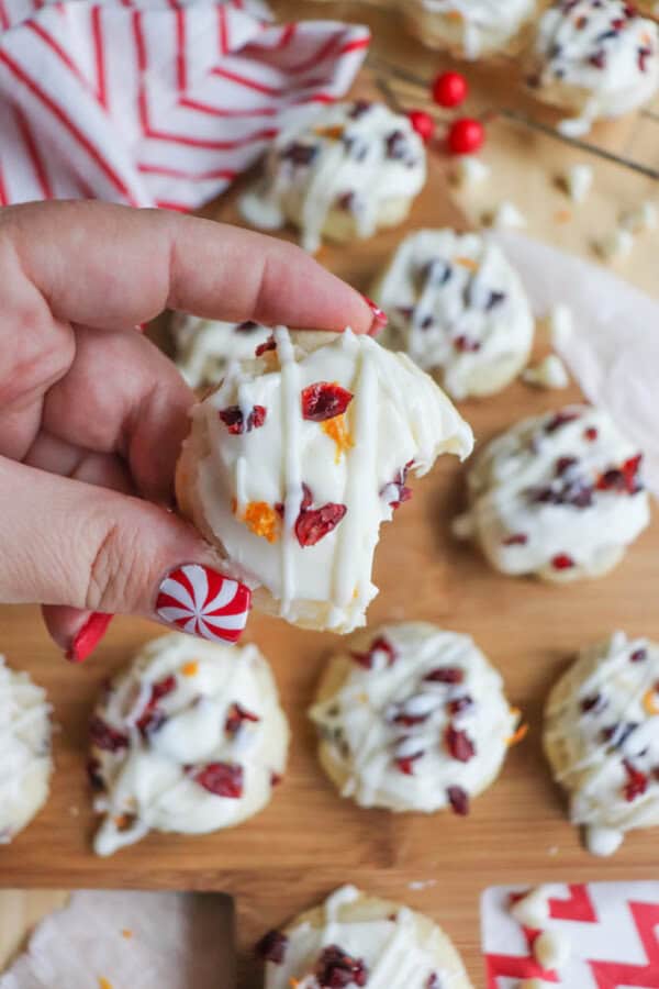A hand holds a cookie topped with white icing, orange zest, and cranberries. The cookie has a bite taken out of it. More decorated cookies are on a wooden surface in the background. The persons nails are painted with a candy cane design.