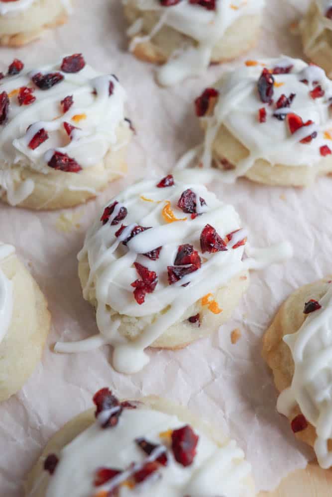 Close-up of cookies topped with white icing and sprinkled with dried cranberries and orange zest, arranged on parchment paper.