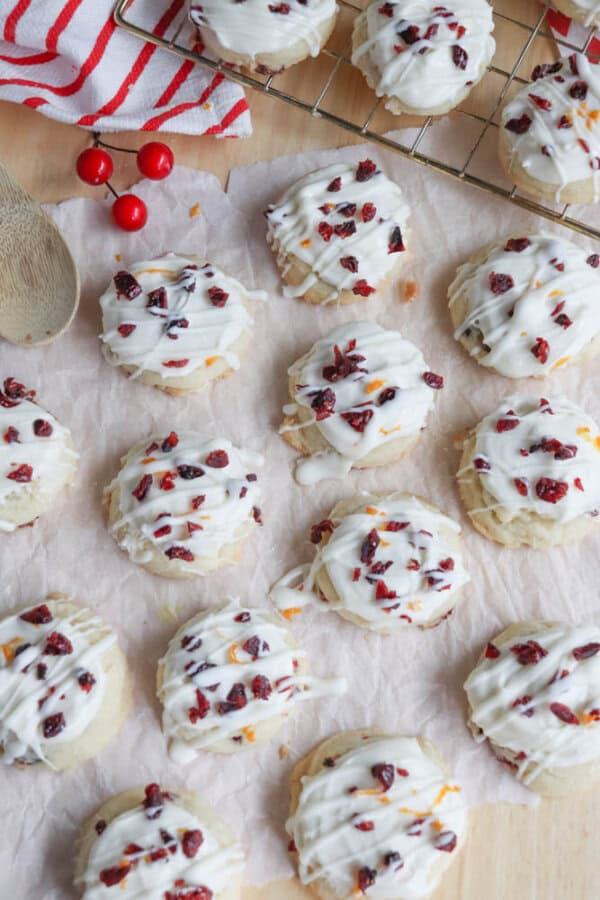 White chocolate cranberry cookies on parchment paper, topped with white chocolate drizzle and dried cranberries, with a striped cloth and cooling rack in the background.