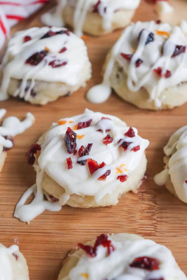 A close-up of cookies on a wooden surface, drizzled with white icing and topped with small pieces of dried cranberries. The icing appears creamy and some of it has dripped onto the surface around the cookies.