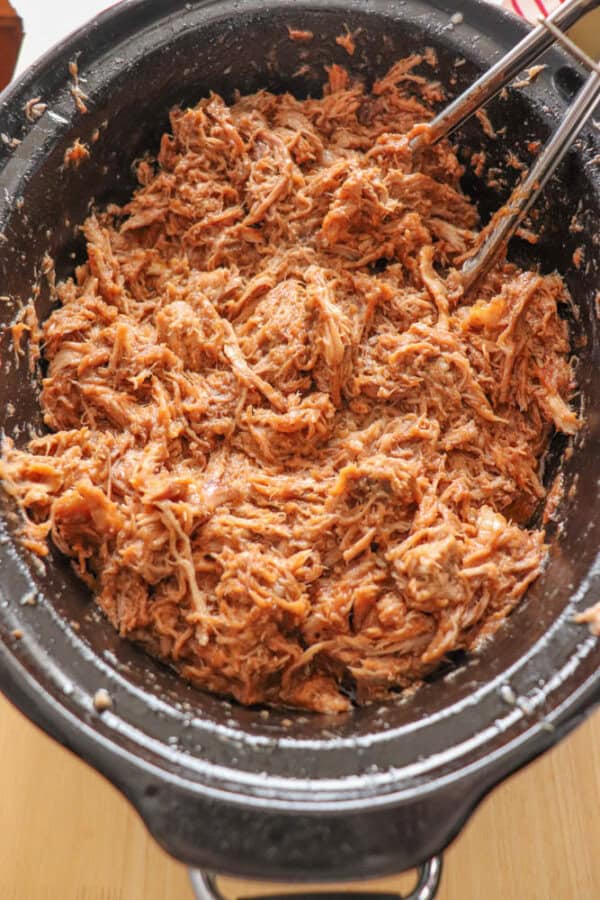 A close-up of crockpot pulled pork in a black slow cooker. Two metal tongs rest beside the tender, seasoned meat, perfectly shredded and ready for serving.