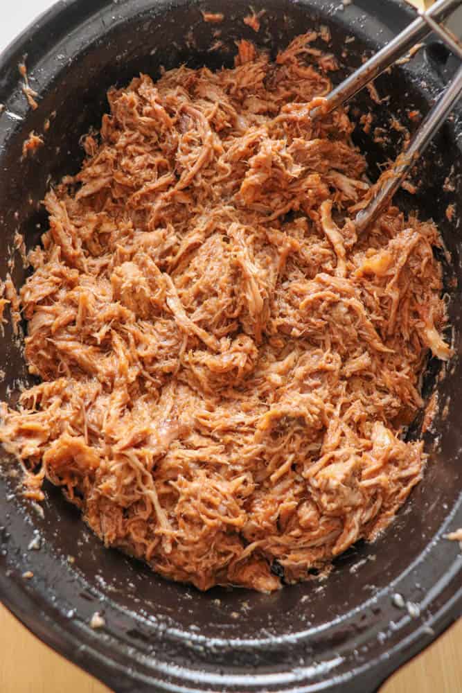 Shredded crockpot pulled pork in a black slow cooker with metal tongs on the side. The pork is well-cooked, tender, and juicy, showcasing a rich blend of light and dark brown hues.