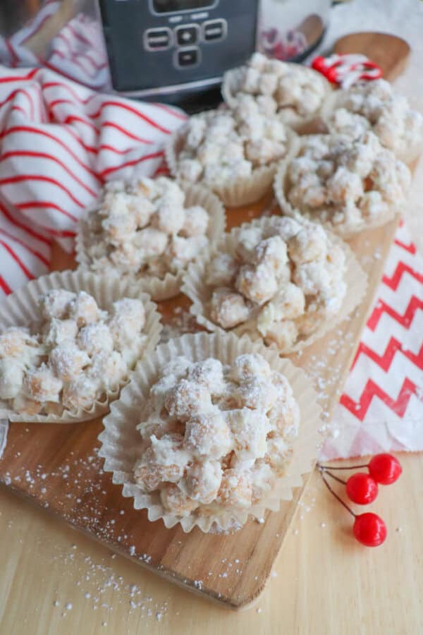 Seven clusters of white, powdered cereal treats are placed in cupcake liners on a wooden board. A striped red and white cloth is in the background, and red cherries are nearby. A black electronic device is partially visible.