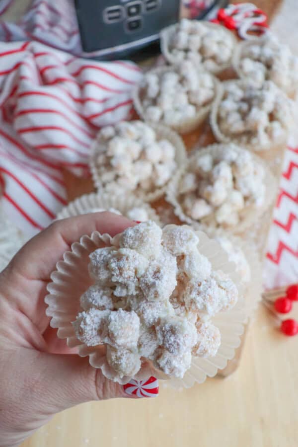 A hand holds a paper liner filled with powdered sugar-coated snack clusters, with more clusters arranged on a wooden board in the background. A red and white striped cloth adds a festive touch.