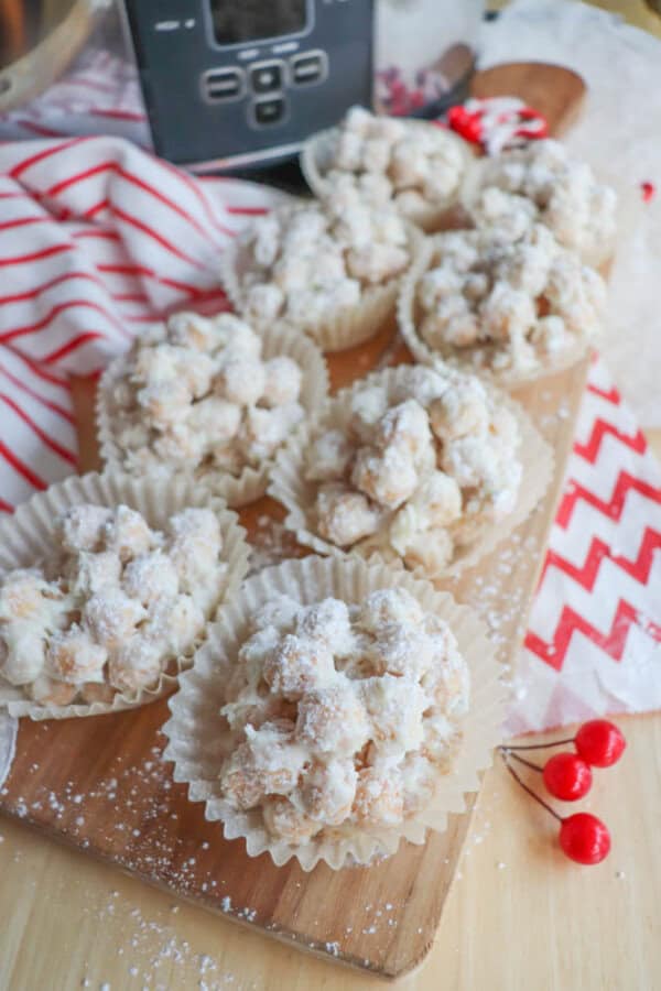 Mini powdered sugar-coated desserts in paper liners are arranged on a wooden board. A red and white striped cloth and a zigzag-patterned napkin are nearby, along with a few red berries. A kitchen appliance is in the background.