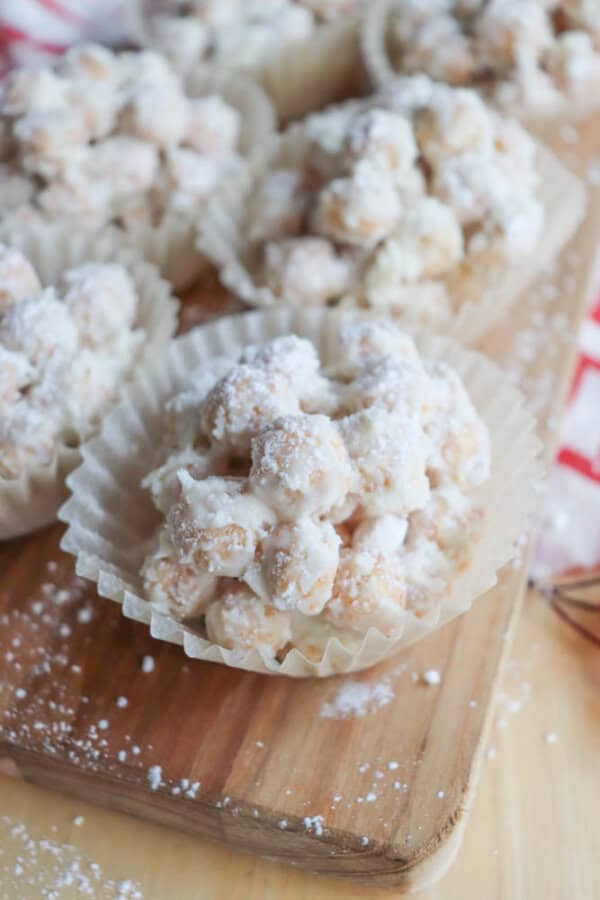 Close-up of clusters of white chocolate-coated popcorn or cereal, arranged in paper baking cups on a wooden board. The treats are dusted with powdered sugar, adding a festive touch.