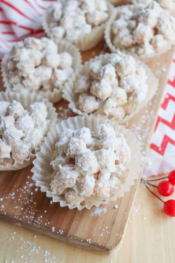 Close-up of several snowball cookies topped with powdered sugar, arranged on a wooden board. Theyre placed in white paper cupcake liners. A red and white cloth and red berries are partially visible in the background.