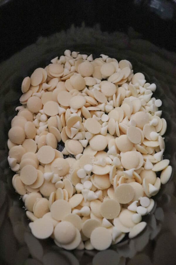 A pile of white chocolate chips and discs in varying sizes is spread inside a dark container. The smooth, creamy texture of the chocolate contrasts with the dark background.