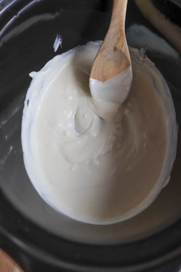 A wooden spoon stirring creamy white chocolate or melted candy inside a black slow cooker. The mixture appears smooth and glossy.