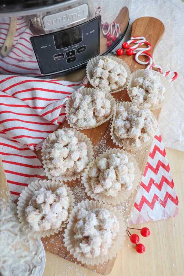 A wooden board holds several clusters of white chocolate-coated cookies in paper cups, dusted with powdered sugar. A red and white striped cloth, candy canes, and red berries are nearby. In the background is a slow cooker.