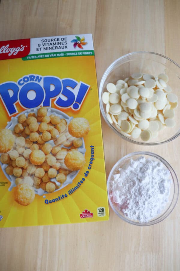 Box of Kelloggs Corn Pops cereal on a wooden surface, next to a bowl of white chocolate chips and a bowl of powdered sugar.