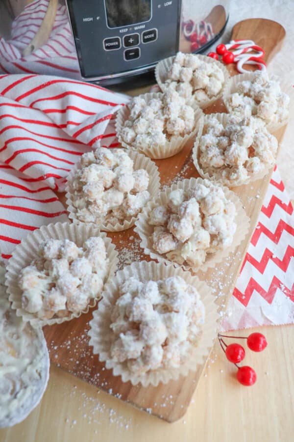 Nine paper cups filled with white chocolate-covered clusters sit on a wooden board. A red and white striped cloth and a red chevron napkin are nearby. Small red decorative berries are scattered around.