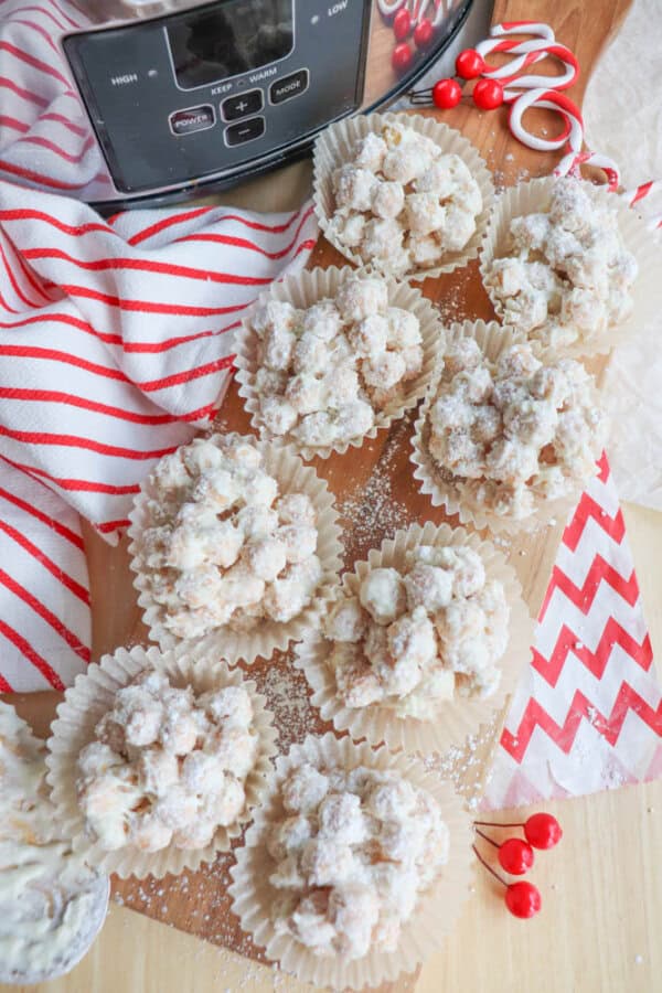 Powdered sugar-covered clusters arranged on a wooden cutting board, with several placed in white cupcake liners. A red and white striped cloth, red berries, and a slow cooker partially visible in the background.