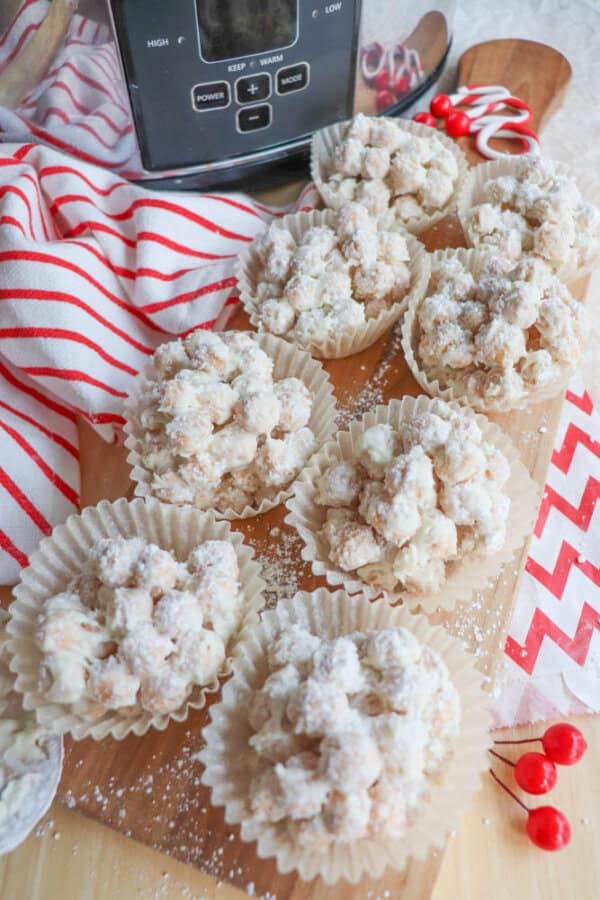 Homemade treats topped with powdered sugar are arranged on a wooden board. They are placed in white cupcake liners, surrounded by a striped red and white cloth, a slow cooker, and decorative red berries.