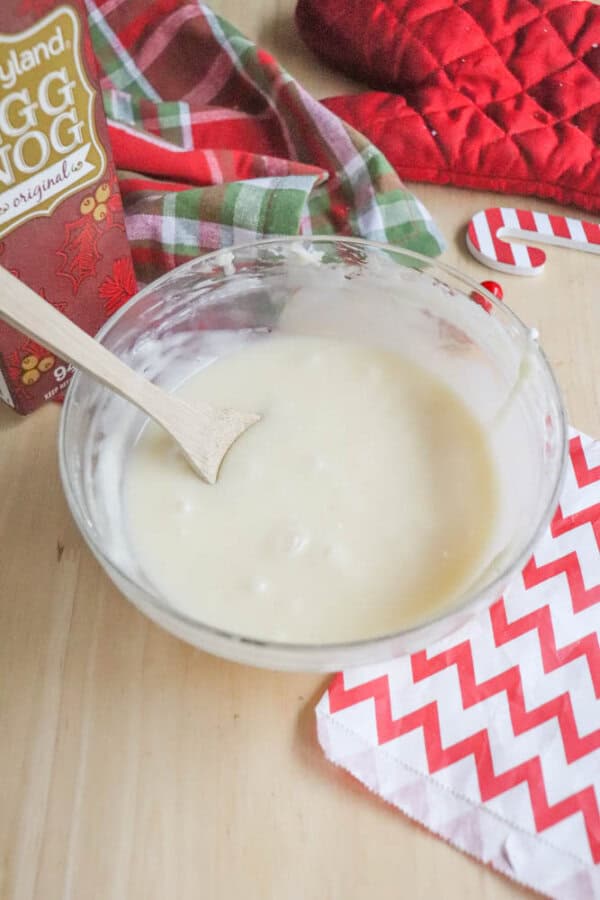 A mixing bowl containing a creamy eggnog icing with a wooden spoon rests on a wooden surface. Nearby are a carton of eggnog, red and green cloth, a red oven mitt, a candy cane, and a red and white chevron-patterned napkin.