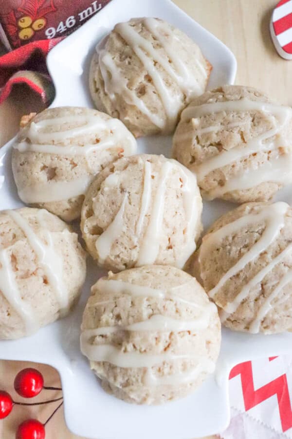 A plate of six round cookies drizzled with icing. The cookies have a crumbly texture and sit on a square, white plate. A red and white chevron napkin and some red decorative berries are partially visible in the background.