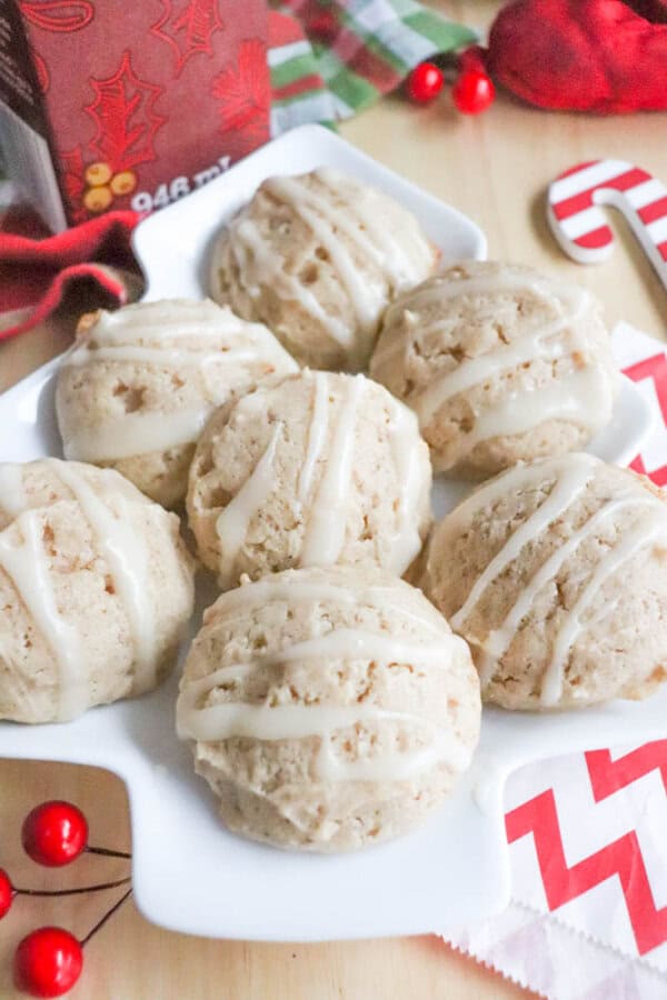 Eggnog Cookie Recipe A white plate with six round, iced cookies. The cookies have a drizzle of white icing and are surrounded by festive decorations, including red berries and candy canes on a light wood table.