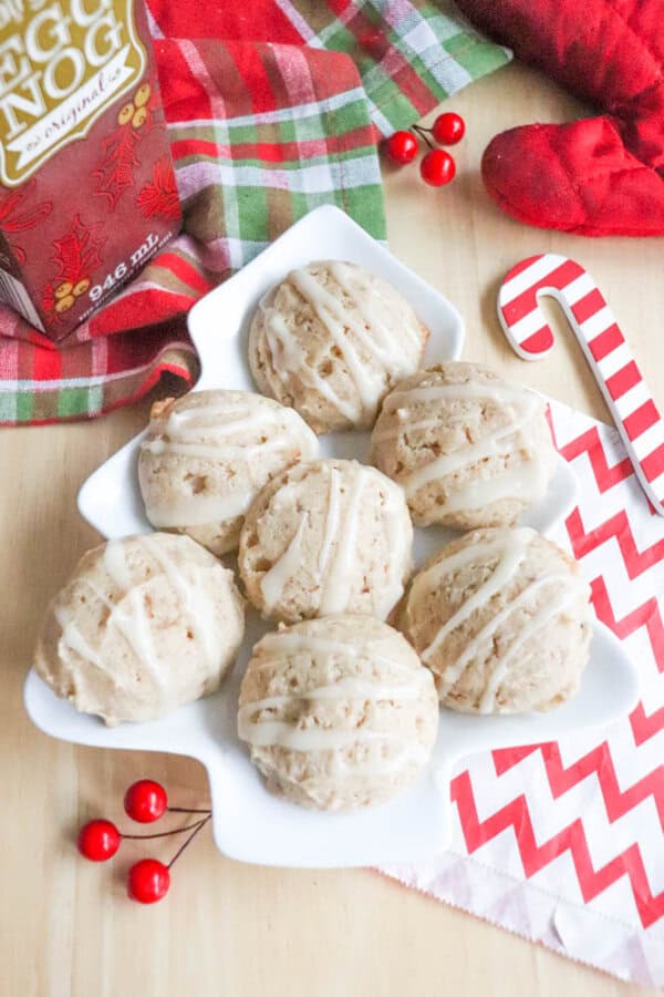 Cookies with drizzled icing arranged on a tree-shaped plate, accompanied by a carton of eggnog, red berries, a red oven mitt, a candy cane, and a red and white chevron napkin.