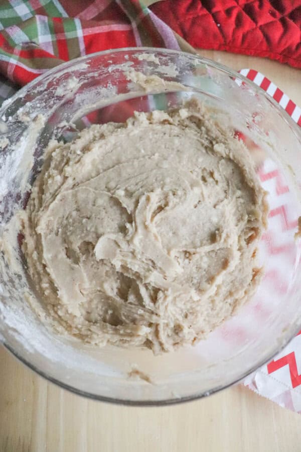 A bowl of cookie dough sits on a wooden surface. The dough is creamy and mixed thoroughly. In the background, there are colorful checkered and red oven mitts.